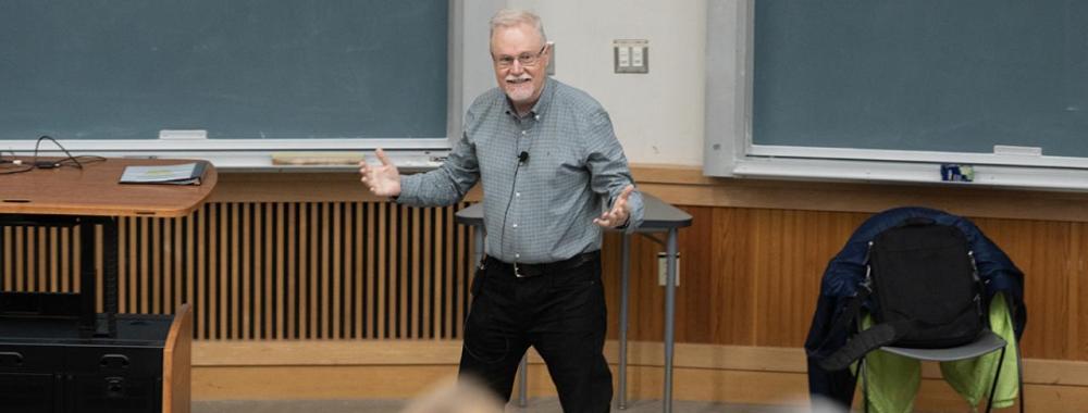 Professor Steven Luck standing in front of a classroom with a clip on mic. His hands are spread wide apart, palms facing up, smiling as he is eagerly lecturing.
