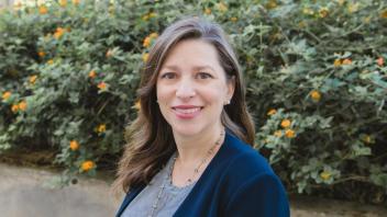 A headshot of Marina Leite outside in front of flowering bushes. She is wearing a blazer with her long hair to the side. 