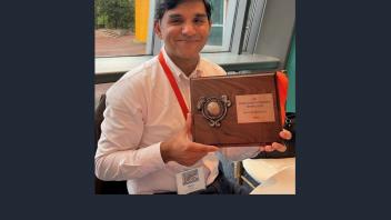 Smiling man in white shirt holding a wooden award plaque indoors by a window
