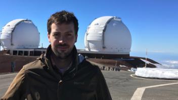 Man in jacket at mountain observatory with two white telescope domes and clear blue sky