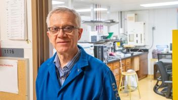 Older man in blue lab coat and glasses, standing in a busy laboratory