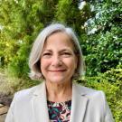 Smiling woman with gray hair, wearing a light blazer, in a green outdoor setting.